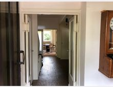 Blick durch einen schmalen Flur in einen Raum mit Tisch und Stühlen vor einem großen Fenster. Rechts an der Wand hängt eine Holz-Wanduhr, links ist eine geöffnete Glastür zu sehen.