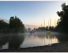 Ein See bei Sonnenaufgang mit Nebel, umgeben von Bäumen. Auf dem Wasser schwimmt eine leuchtende Lotusblüten-Skulptur, und im Hintergrund ragen mehrere hohe Funkmasten in den Himmel.