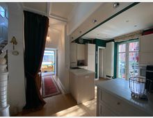 Helle, moderne Innenansicht einer Küche mit weißer Kücheninsel, hohen Decken und französischen Doppeltüren, die auf eine Terrasse führen. Links befindet sich ein Eingangsbereich mit einem roten Teppich und einer Treppe, die nach oben führt.