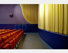 Innenansicht eines kleinen Kinos mit orangefarbenen Polstersitzen, einer lila und blauen Wandgestaltung sowie einem geschlossenen gelben Vorhang vor der Leinwand. Der Boden besteht aus grauem Material, und an der Seitenwand ist ein Lautsprecher zu sehen.