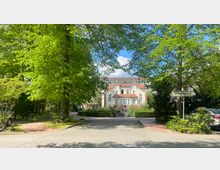 Historisches weißes Gebäude mit roten Dachziegeln, umgeben von Bäumen und gepflegten Gärten; im Vordergrund eine gepflasterte Einfahrt, rechts ein Schild mit der Aufschrift "Ausfahrt".