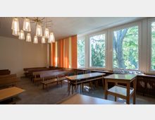 Gerichtssaal mit schlichten Holzbänken und -tischen, großen Fenstern mit Blick auf Bäume und orange-gelben Wandpaneelen. Eine auffällige, moderne Deckenleuchte hängt von der weißen Decke.