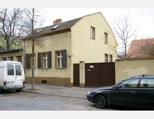 Zweistöckiges Wohnhaus mit gelber Putzfassade, dunklen Fensterläden und Satteldach, aufgenommen in einer ruhigen Straße mit Kopfsteinpflaster. Vor dem Gebäude befinden sich geparkte Fahrzeuge und ein braunes Tor an einer angrenzenden Mauer.