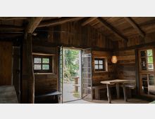 Holzhaus-Interieur mit rustikaler Einrichtung: Ein Raum aus Holz mit unverputzten Wänden und einer offenen Tür, die nach draußen führt. Innen befinden sich kleine Fenster, eine einfache Sitzbank, ein Holztisch, und eine Hängelampe, die warmes Licht spendet.