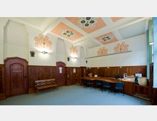 Ein historischer Büroraum mit Holztäfelung an den Wänden, verzierten Holztüren und dekorativ bemalten Deckenpanelen in Orange- und Weißtönen. Ein langer Schreibtisch mit mehreren Stühlen und Computerarbeitsplätzen dominiert den Raum, der von hohen Fenstern mit Vorhängen beleuchtet wird.