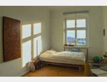 Helles Schlafzimmer mit Holzboden, einem schlichten Bett auf Paletten und einem großen Fenster, durch das Tageslicht fällt. Neben dem Bett stehen kleine Regalelemente mit persönlichen Gegenständen, und an der Wand hängt eine dekorative Holztafel.