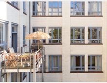 Großes Wohngebäude mit hellen Ziegelwänden und blauen Fensterrahmen; im Vordergrund ein kleiner Balkon mit Holzstühlen, Blumentöpfen und einem Sonnenschirm aus Strohmaterial.