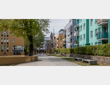 Wohngebiet mit bunten Mehrfamilienhäusern, Gehweg und Grünflächen; links im Hintergrund eine Kirche mit hoher Spitze, daneben moderne Gebäude aus Backstein und Glas.