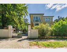 Zweistöckige Stadtvilla mit gelber Putzfassade, weißen Gesimsen und einem kleinen Balkon über dem Eingang, umgeben von Bäumen und Sträuchern. Vor dem Haus stehen eine niedrige Mauer und ein schmiedeeisernes Tor mit Zufahrt von einer gepflasterten Straße.