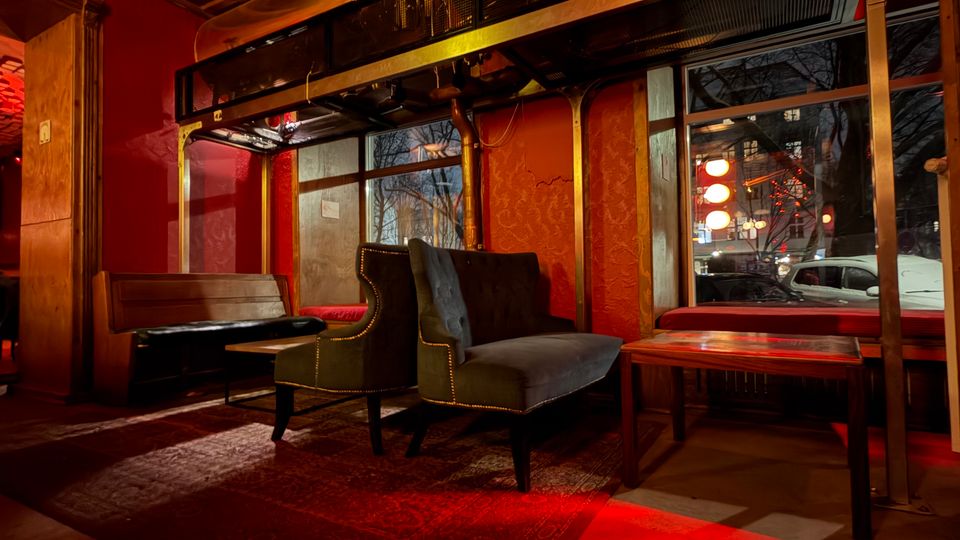 Innenraum einer Bar oder Lounge mit roten Wänden, gemustertem Teppich und dunklen Sitzbänken sowie Sesseln um niedrige Tische. Über den Fenstern verläuft eine goldfarbene Metallkonstruktion unter einer verzierten Kassettendecke, mit warmem, rötlichem Licht und Blick nach draußen auf die Straße.