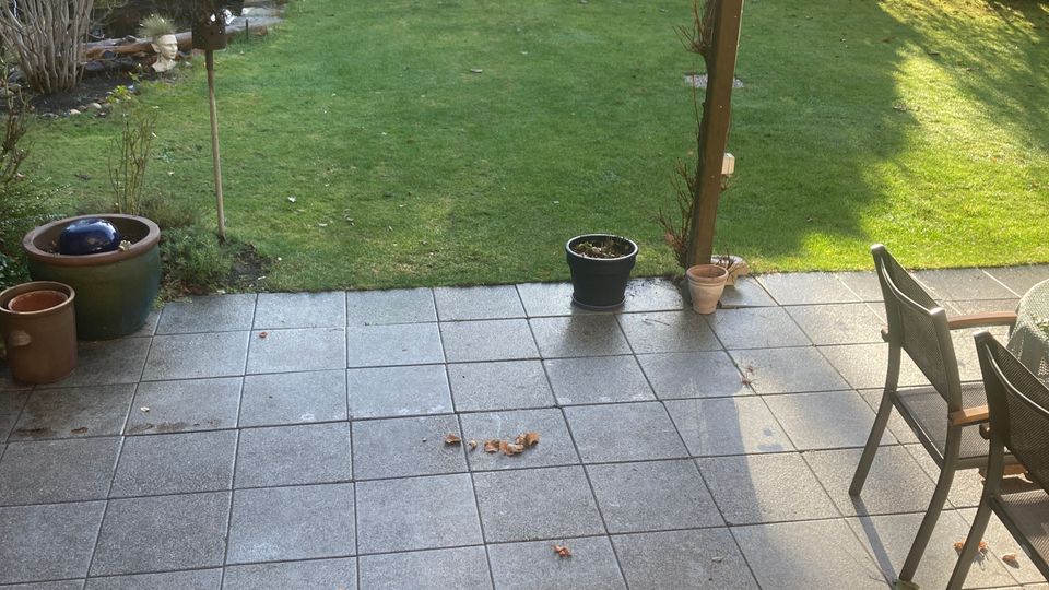 Eine überdachte Terrasse mit Fliesenboden und Holzstufen, die in einen Garten führt. Der Garten hat eine Rasenfläche, umgeben von Büschen und kleinen Dekorationen, wie hängende Ornamente und Blumentöpfe.
