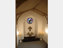 Innenraum einer kleinen Kapelle mit schlichtem Altar aus Stein, einer kreuzförmigen Wandverzierung und einem runden Buntglasfenster darüber. Auf dem Altar stehen Kerzen und Blumengestecke, daneben befinden sich zwei Kerzenständer mit mehreren angezündeten Kerzen.