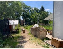 Ein großzügiger Garten mit verschiedenen Elementen: links ein kleines Gebäude mit Wellblechdach und ein Trampolin, in der Mitte ein Weg aus Holzschnitzeln, rechts eine alte Backsteinmauer mit integriertem Pizzaofen. Im Hintergrund sind ein Spielplatz und dichte Bäume zu sehen.