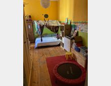 Kinderzimmer mit gelben Wänden und Holzboden, eingerichtet mit einem Bett, das mit einem dekorativen Stoffbaldachin versehen ist. Im Raum befinden sich außerdem ein roter Teppich, ein kleines Trampolin, ein gelber Hocker, Regale mit Spielzeug und Büchern sowie diverse Kinderzeichnungen an der Wand.