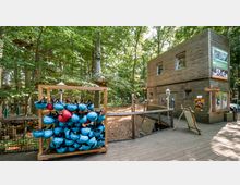 Holzkonstruktionen in einem Kletterpark inmitten eines Waldes, mit einem Gebäude aus Holzpaneelen im rechten Bildbereich und einem Regal mit blauen und roten Helmen im Vordergrund. Im Hintergrund sind zwischen den Bäumen Plattformen und Seilelemente erkennbar.