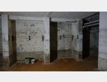 Kellerraum mit Sichtmauerwerk und abblätterndem Putz, unterteilt durch Betonpfeiler. Auf dem Boden stehen ein grüner Eimer und ein weißer Farbeimer, während an der Decke Rohre und Kabel verlaufen.