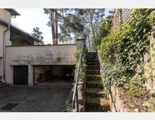 Ein Außenbereich mit einer Garage, die teilweise geöffnet ist und Fahrräder sowie ein Auto darin zeigt. Rechts daneben eine steile Treppe aus Stein mit einem Metallgeländer, umgeben von bewachsenen Mauern und Efeupflanzen.