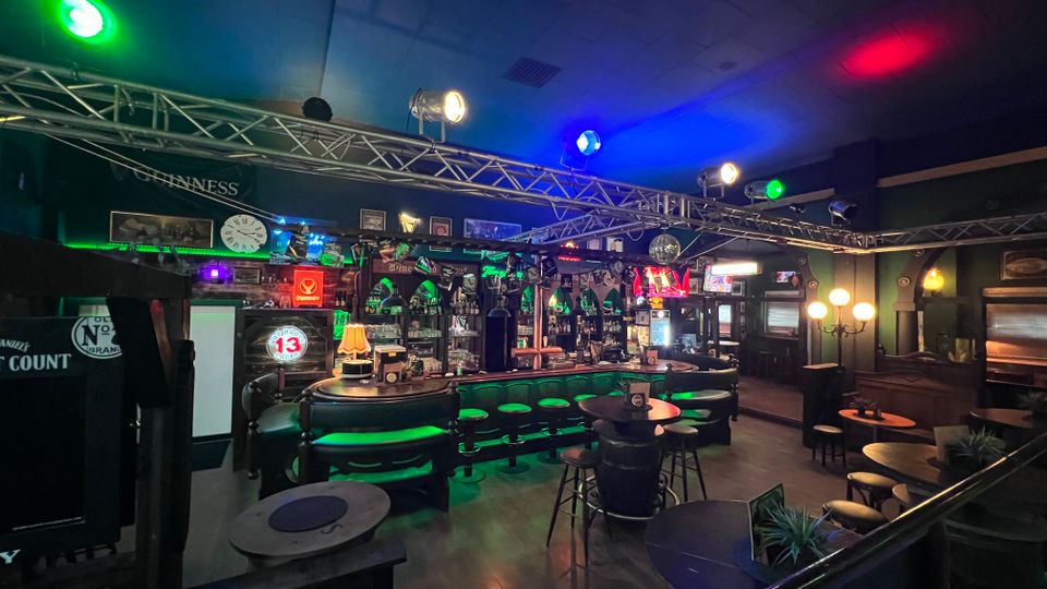 Innenraum einer dunklen Bar mit geschwungener Theke, beleuchteten Barhockern und vielen Spirituosenflaschen im Regal. Über der Sitzfläche hängen Traversen mit farbigen Bühnenlichtern; im Raum stehen mehrere kleine Rundtische und Sitznischen an den Wänden.