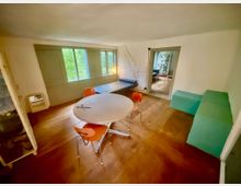 Heller Wohnraum mit breiten Holzdielen, weiß verputzten Wänden und großen Fenstern mit grünen Klappläden. In der Mitte stehen ein runder Tisch mit orangefarbenen Stühlen, dazu eine Liege an der Fensterwand und türkisfarbene Sitzblöcke neben einer offenen Tür zu einem Nebenraum.