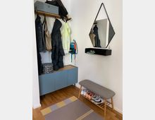 Eingangsbereich mit einer blauen Kommode, einer Holz-Garderobenleiste mit Jacken und Taschen, einem Wandspiegel in geometrischer Form und einem kleinen schwarzen Wandregal. Rechts steht eine gepolsterte Holzbank mit Schuhen darunter, der Boden besteht aus Holz und liegt ein bunter Teppich.