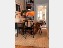 Esszimmer in einer Wohnung mit Holzboden und einem runden Esstisch, der von Stühlen in unterschiedlichen Designs umgeben ist. Im Hintergrund sind ein Regal mit Büchern und Deko sowie ein angrenzendes Zimmer mit einem Bett und großen Fenstern zu sehen.