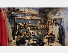 Werkstatt für Schuhmacher mit einer alten Nähmaschine im Vordergrund, Lederschuhen, Werkzeugen und Materialien auf Regalen und an den Wänden. Eine Schaufensterpuppe trägt Arbeitskleidung und sitzt am Tisch, umgeben von verschiedenen Schuhen und Handwerksutensilien in einem rustikalen, industriellen Raum.