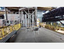 Industrielle Werkshalle mit metallischen Rohrleitungen und Geländern; im Vordergrund ein kleiner Sitzbereich mit einem Tisch und vier Stühlen, umgeben von gelben Sicherheitsgittern. Im Hintergrund sind Maschinen, Stahlträger und aufgerollte Kabel erkennbar.