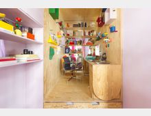 Narrow built-in study nook finished in light plywood, with a small desk, shelves, and wall-mounted cubbies filled with books and colorful objects. A black swivel chair sits in the center, and the space is integrated into a room with pale pink shelving on the left.