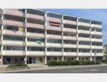 Mehrstöckiges Wohngebäude mit Balkonen, von denen einige Markisen oder Dekorationen aufweisen. Im Erdgeschoss befinden sich Geschäfte und ein Eingang, davor sind eine kleine Grünanlage sowie ein Gehweg mit Straßenzugang.