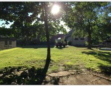 Grasbewachsene Fläche mit vereinzelten Bäumen und Gebäuden im Hintergrund, darunter ein graues Gebäude mit Fenstern. Im Vordergrund verläuft ein schmaler gepflasterter Weg, und in der Mitte des Bildes steht ein kleiner roter Traktor. Die Sonne scheint durch die Bäume.