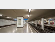 Ein moderner U-Bahnhof mit gekachelten Wänden, rechteckigen weißen Säulen und mehreren Bahnsteigen. Auf einer Wand ist der Schriftzug "Potsdamer Platz" zu lesen, und beleuchtete Gleisanzeigen hängen von der Decke.