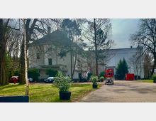 Weißes Gebäude mit Walmdach und einem Anbau, umgeben von Bäumen und einer gepflasterten Zufahrtsstraße. Im Vordergrund stehen Pflanzen in Töpfen, parkende Autos und ein roter Hebelift, rechts ein roter Container.