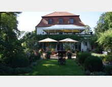 Zweigeschossiges Haus mit rotem Satteldach und weißer Fassade, umgeben von einem üppigen Garten mit Rasen, Hecken und blühenden Pflanzen. Vor dem Haus befinden sich eine Terrasse mit Sonnenschirmen, Sitzmöbeln und einem Gartenofen, teilweise von Efeu umrankt.
