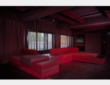 Dunkler Innenraum mit niedriger Sitzgruppe aus roten Polstern, die in einer Eckform angeordnet ist. Im Hintergrund befinden sich große Fenster mit vertikalen Metallstangen als Raumteiler und eine offene Decke mit sichtbaren Rohren.