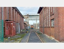 Schmaler Bahngleisabschnitt zwischen zwei langen, roten Backsteingebäuden eines Industriestandorts, mit einer kleinen orangefarbenen Lokomotive im Hintergrund und einem Metallsteg, der die Gebäude verbindet. Im Vordergrund sind Absperrungen, Gras und ein Reparaturbereich zu sehen.