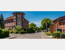 Straßenecke mit einem mehrstöckigen historischen Backsteingebäude mit Turmaufsatz und Rundbogenfenstern, umgeben von Hecken und Bäumen. Rechts steht ein weiteres Backsteingebäude entlang einer ruhigen Straße unter blauem Himmel.