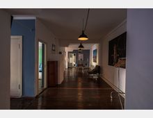 Langer Flur in einem Innenraum mit Holzboden, einer schlichten weißen und blauen Farbgestaltung, und einer Hängelampenbeleuchtung. An den Wänden befinden sich Kunstwerke, Möbel wie Stühle, ein Tisch, und ein rollbarer Garderobenständer.