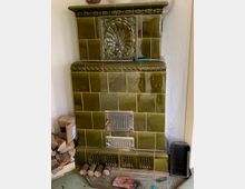 Grüner Kachelofen mit glänzenden Keramikfliesen und dekorativen Verzierungen, darunter ein großes Ornament in Muschelform. Vor dem Ofen liegen Holzscheite und Briketts, daneben steht ein Holzkorb; im Hintergrund ist eine helle Wand sichtbar.