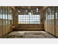Ein verlassenes, heruntergekommenes Gebäude mit großen, vergitterten Fenstern, teilweise abblätternder Farbe und beschädigten Wänden. Der Raum zeigt alte Rohre unter dem Fenster sowie einen kleinen Waschbecken an der linken Wand, mit Schutt und Peeling-Farbe auf dem Boden.
