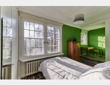 Schlafzimmer mit großen Fenstern und Blick ins Freie, grün gestrichenen Wänden und Holzfußboden. Im Raum befinden sich ein Bett mit kariertem Bettbezug, ein Holzschrank, ein Schreibtisch mit Stuhl sowie eine Stehlampe mit bunten Lampenschirmen.