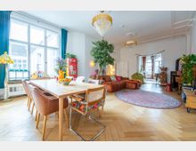 Großzügiger, gemütlicher Wohn- und Essbereich in einer hellen Wohnung mit Parkettboden. Der Raum enthält einen Esstisch mit Stühlen, eine große Fensterfront mit blauen Vorhängen, eine braune Sofalandschaft, mehrere Zimmerpflanzen und dekorative Kronleuchter.