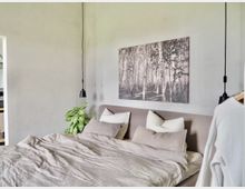 Schlafzimmer mit einem großen, grauen Bett, das mit beigen und weißen Kissen sowie einer passenden Decke dekoriert ist. Im Hintergrund eine graue Wand mit einem Wandbild eines Birkenwaldes und zwei schwarzen, herabhängenden Hängeleuchten; links eine Zimmerpflanze.