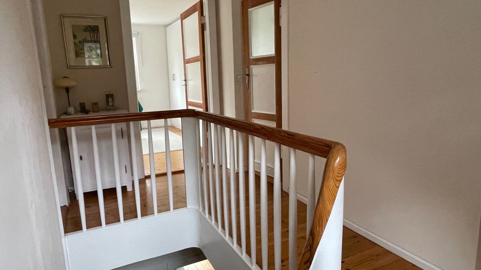 Flur im Obergeschoss eines Wohnhauses mit Holzfußboden und Treppengeländer aus weißen Sprossen mit Holzhandlauf. Mehrere Türen mit Holzrahmen und Milchglasscheiben führen in angrenzende Räume; an der Wand steht eine Kommode mit Lampe und Bildern.