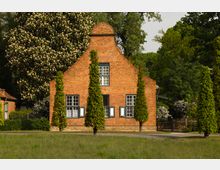 Backsteinhaus mit Satteldach und drei symmetrischen Fenstern an der Fassade, umgeben von hohen, kegelförmig geschnittenen Bäumen und grünem Rasen, im Hintergrund dichte Vegetation und blühende Sträucher.