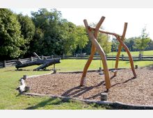 Spielplatz in einer natürlichen Umgebung mit Holzschaukeln und einem holzgesäumten Bereich mit Rindenmulch. Im Hintergrund befinden sich Wiesen, Bäume und ein Holzzaun.
