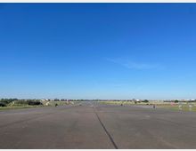 Eine weitläufige asphaltierte Fläche, die ehemals als Fluglandebahn genutzt wurde, mit einigen Radfahrern und Spaziergängern in der Ferne. Im Hintergrund sind grüne Wiesen, kleine Bäume und eine städtische Skyline unter klarem, blauem Himmel zu sehen.