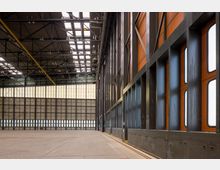 Große Industriehalle mit hoher Decke, sichtbaren Stahlträgern und teilweise verglasten Fassaden. Die seitlichen Wände zeigen Fensterreihen und orangefarbene Paneele, während der Boden aus Beton besteht.