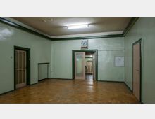 Ein leerer Flur in einem älteren Gebäude mit grünen Wänden, einer Holzbodenfläche und Türen mit grünen Rahmen. Über der zentralen Doppeltür aus Glas hängt eine analoge Wanduhr, im Hintergrund ist ein Treppenhaus sichtbar.