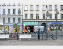 Straßenansicht mit mehrstöckigen Gebäuden, Geschäften im Erdgeschoss, darunter ein Mobilfunkladen, ein Geschäft mit Markise und bunte Kleidung sowie eine Bankfiliale. Im Vordergrund sind ein Telefonhäuschen, zwei weiße Bänke, Fahrräder und Straßenlaternen auf einem gepflasterten Platz zu sehen.