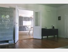 Wohnraum mit Holzboden und einem alten Kachelofen links; durch eine geöffnete Tür ist eine moderne Küche mit grauen Schränken und einem Esstisch zu sehen. Rechts steht eine dunkle Kommode mit einer Tischlampe und Dekorationen.